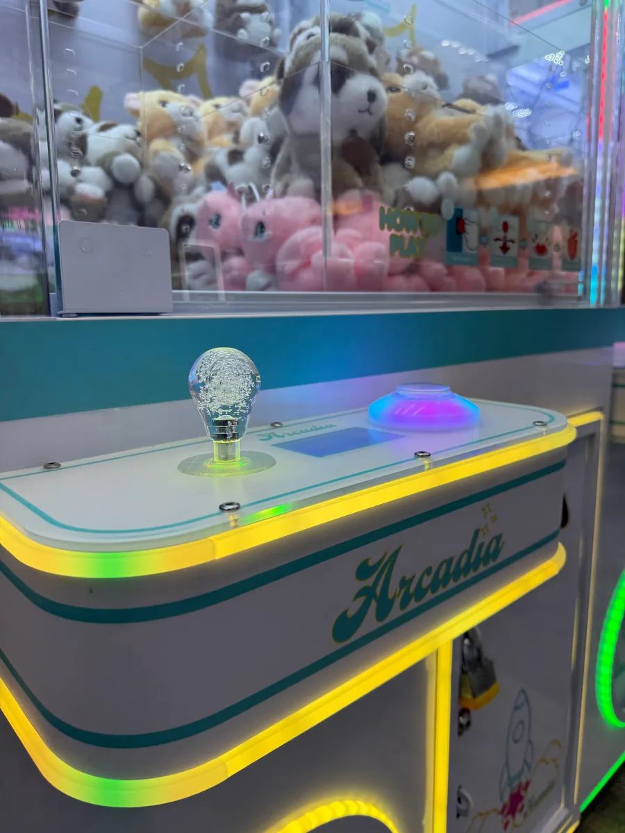 Close-up of an Arcadia claw machine joystick, glowing play button, and plush prizes